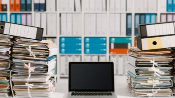 Arbeitsplatz mit vielen Akten links und rechts, in der Mitte ein offener Laptop